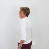An older woman with short brown hair is shown in profile against a plain white wall, wearing a white three-quarter-sleeve top and dark burgundy pants.