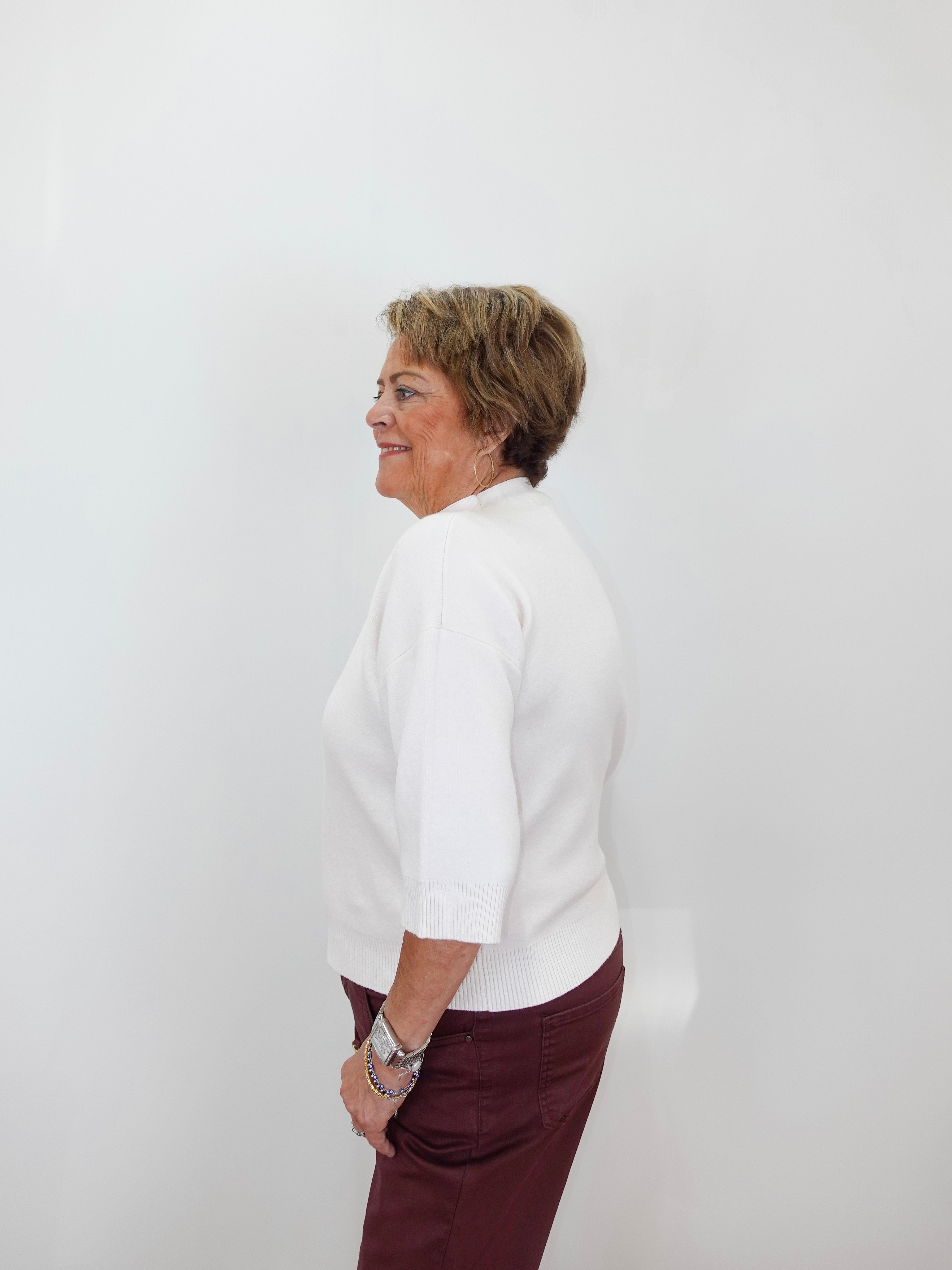 An older woman with short brown hair is shown in profile against a plain white wall, wearing a white three-quarter-sleeve top and dark burgundy pants.