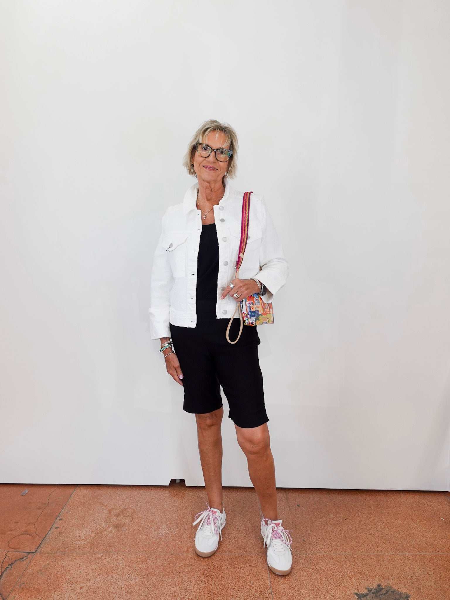 Woman wearing a white jacket, black dress, and shorts with white sneakers against a plain background.