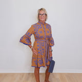 Woman wearing a patterned dress standing against a white wall.