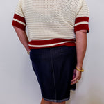 Person wearing a cream sweater with red stripes and a dark denim skirt against a white background