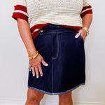 Person wearing a white sweater with red trim and a navy denim skirt against a plain background