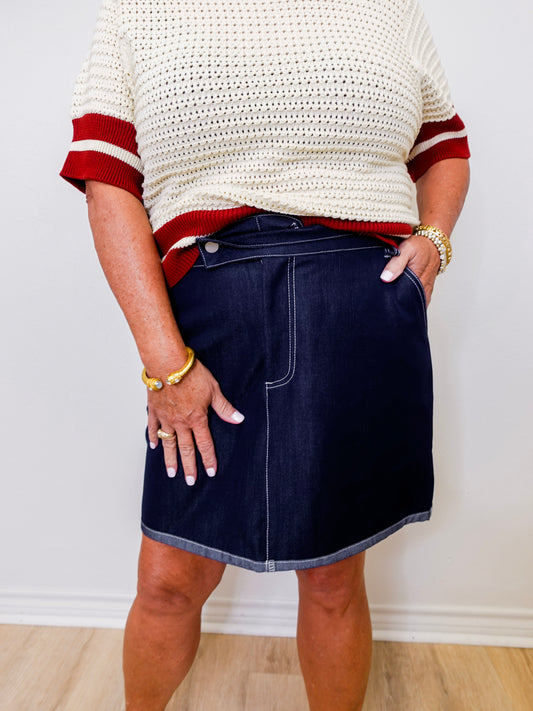 Person wearing a white sweater with red trim and a navy denim skirt against a plain background