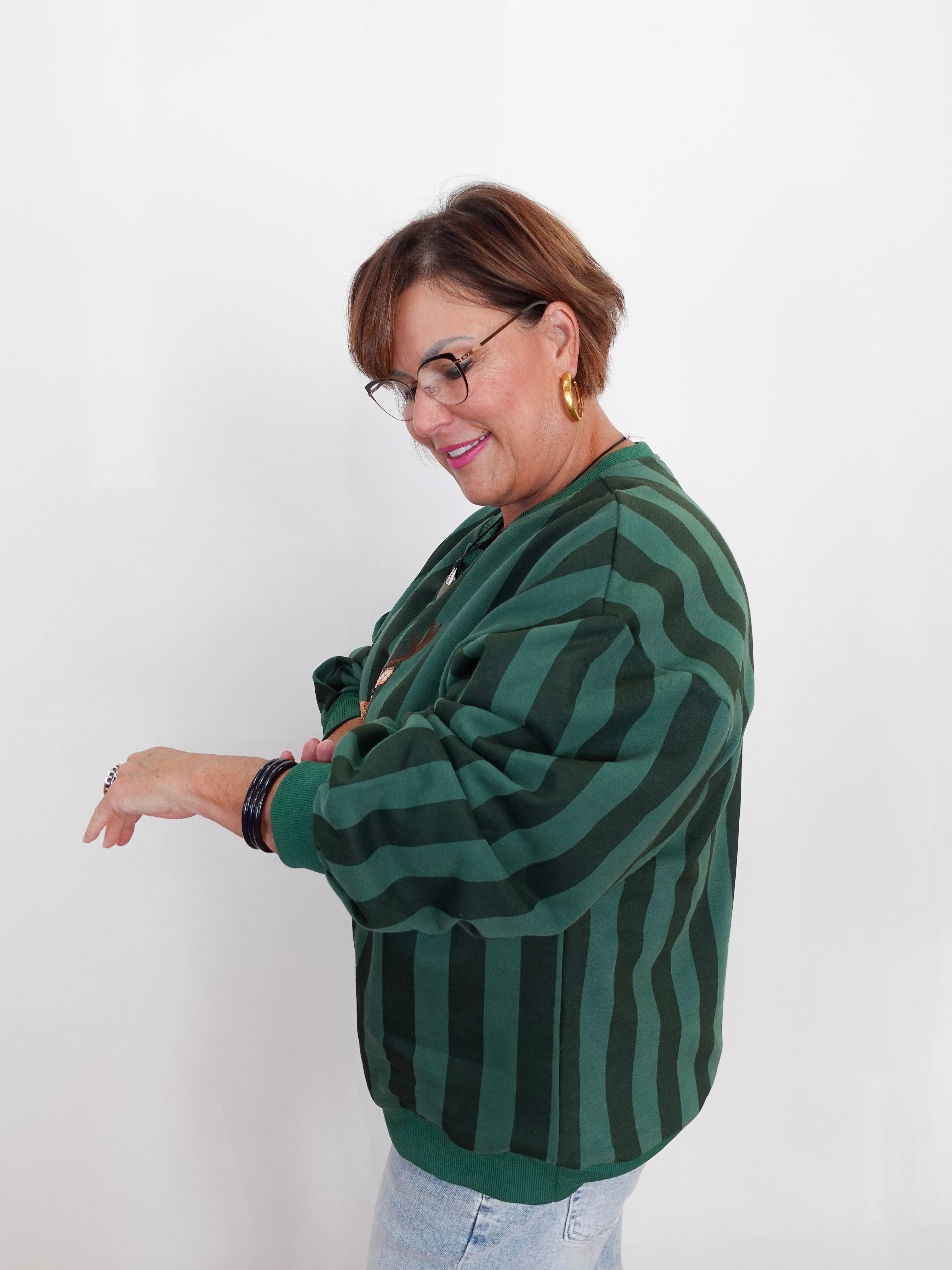 A woman with short brown hair and glasses wearing a green and black vertically striped knit sweater, standing against a plain white background.