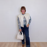 Person wearing a cream vest over a patterned shirt, dark jeans, and brown shoes, holding a white handbag against a plain background.