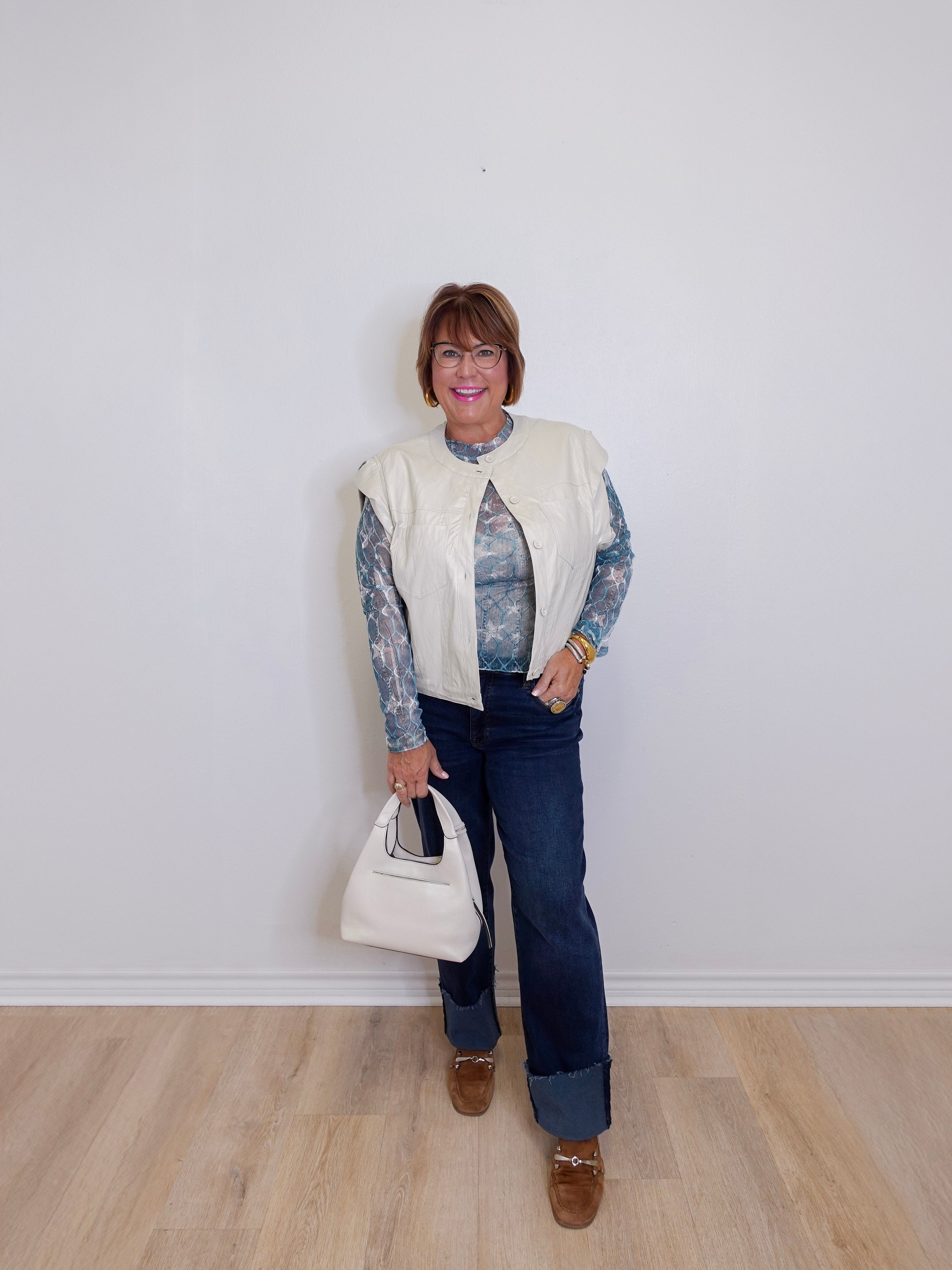 Person wearing a cream vest over a patterned shirt, dark jeans, and brown shoes, holding a white handbag against a plain background.