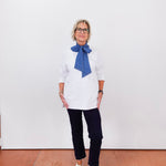 Person wearing a white shirt with a blue scarf and dark pants against a white wall.