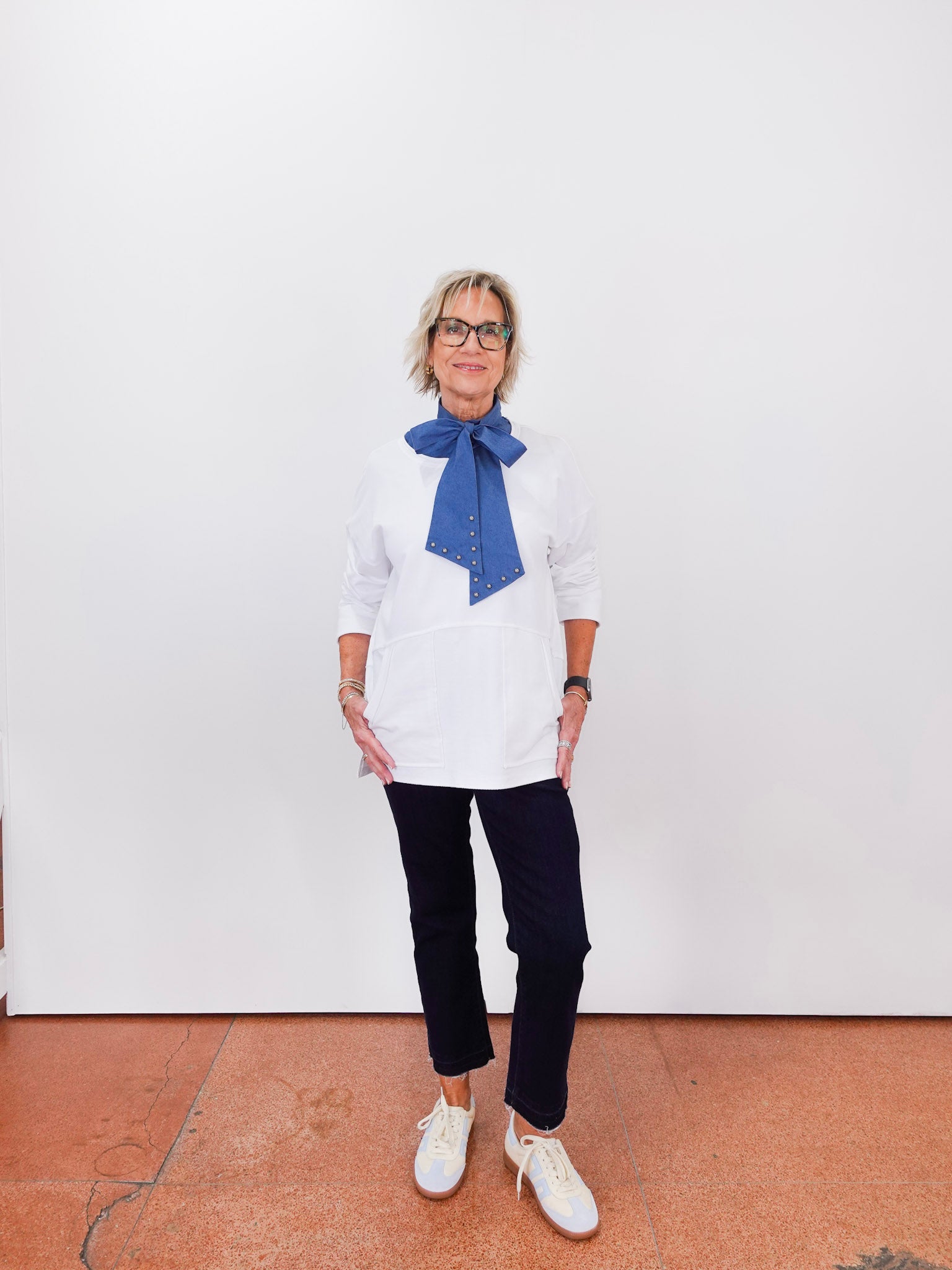 Person wearing a white shirt with a blue scarf and dark pants against a white wall.