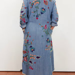 A person viewed from the back wearing a long light blue denim-style embroidered coat or dress with colorful floral and paisley embroidery, standing against a plain white wall.