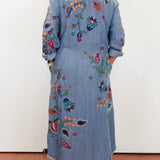 A person viewed from the back wearing a long light blue denim-style embroidered coat or dress with colorful floral and paisley embroidery, standing against a plain white wall.
