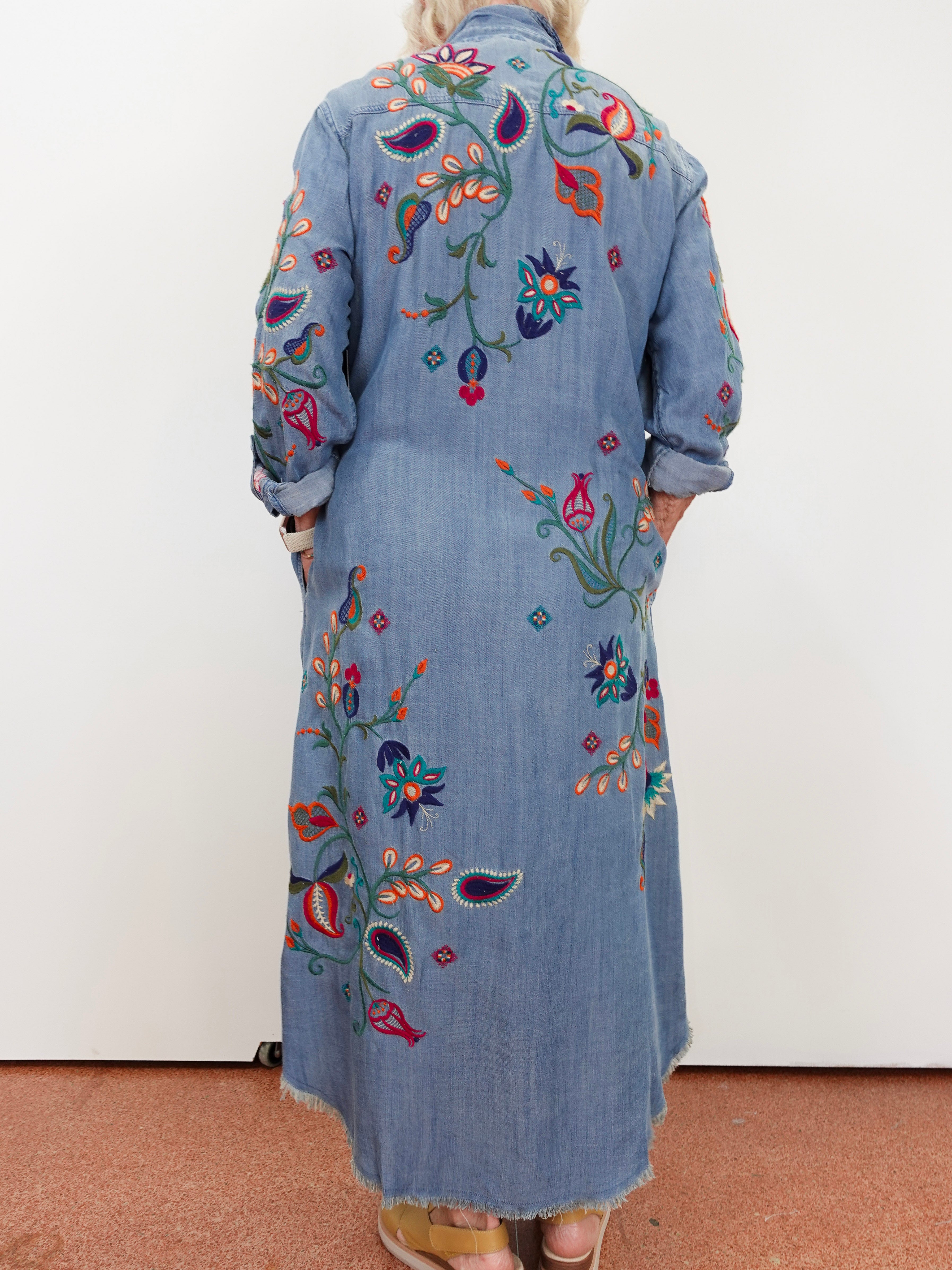 A person viewed from the back wearing a long light blue denim-style embroidered coat or dress with colorful floral and paisley embroidery, standing against a plain white wall.