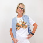 Woman wearing a white t-shirt with a leopard print design, denim vest, and white pants against a plain background.