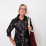 Woman wearing a black patterned shirt and holding a brown bag against a white wall