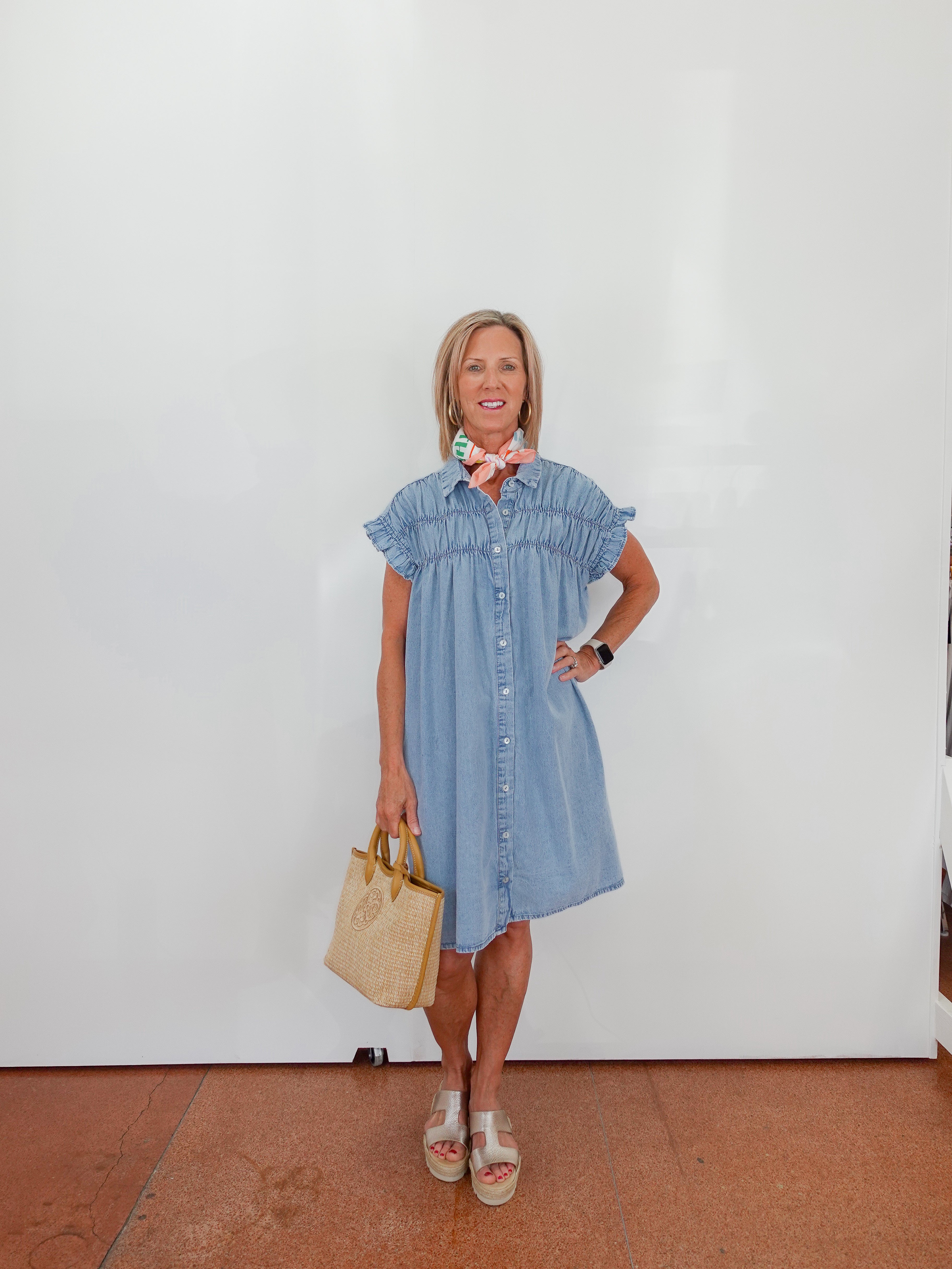Woman wearing a blue dress holding a beige handbag against a white wall.