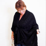 Woman wearing a black poncho and blue jeans, holding a beige handbag indoors.