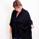 Woman wearing a black poncho and blue jeans, holding a beige handbag indoors.