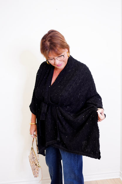 Woman wearing a black poncho and blue jeans, holding a beige handbag indoors.