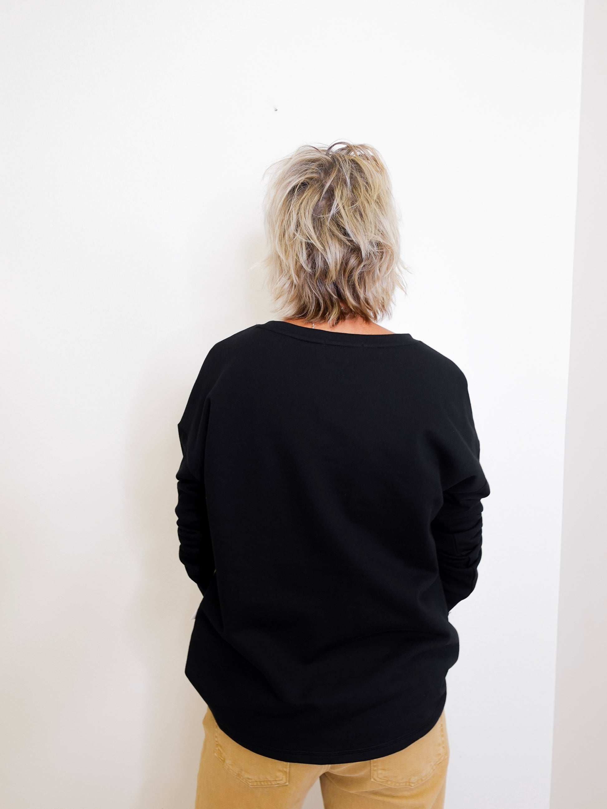 Person wearing a black sweater and beige pants against a white wall