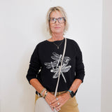 Woman wearing a black sweater with dragonfly design, holding a white bag against a white background