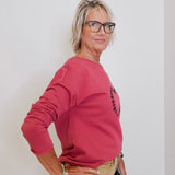 Woman wearing a pink sweatshirt with a logo, standing against a white wall.