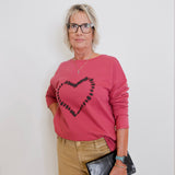 Woman wearing a pink sweater with a heart design, beige pants, and holding a black clutch against a white background.