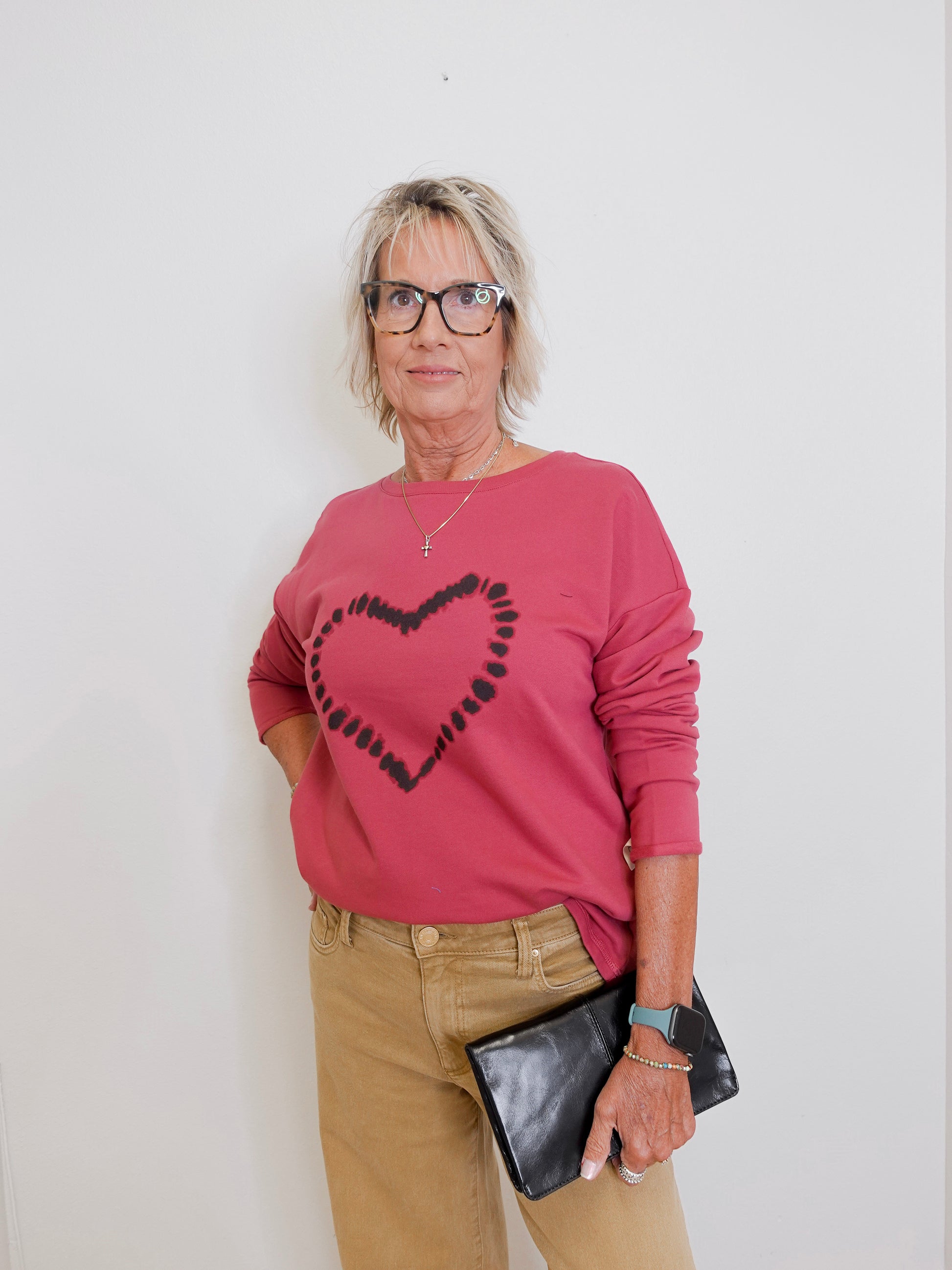Woman wearing a pink sweater with a heart design, beige pants, and holding a black clutch against a white background.