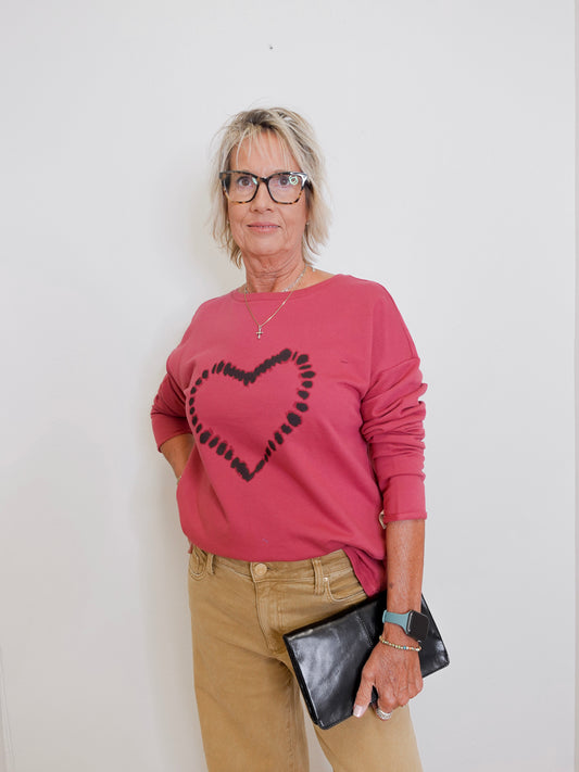Woman wearing a pink sweater with a heart design, beige pants, and holding a black clutch against a white background.