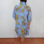 A woman viewed from behind wearing a light blue frayed hem shirt dress with a colorful dragon print, standing against a white wall on a terracotta tiled floor.
