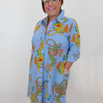 A woman wearing glasses and a light blue collared shirt dress with a colorful dragon print, standing against a plain white wall.