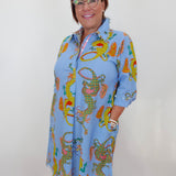 A woman wearing glasses and a light blue collared shirt dress with a colorful dragon print, standing against a plain white wall.