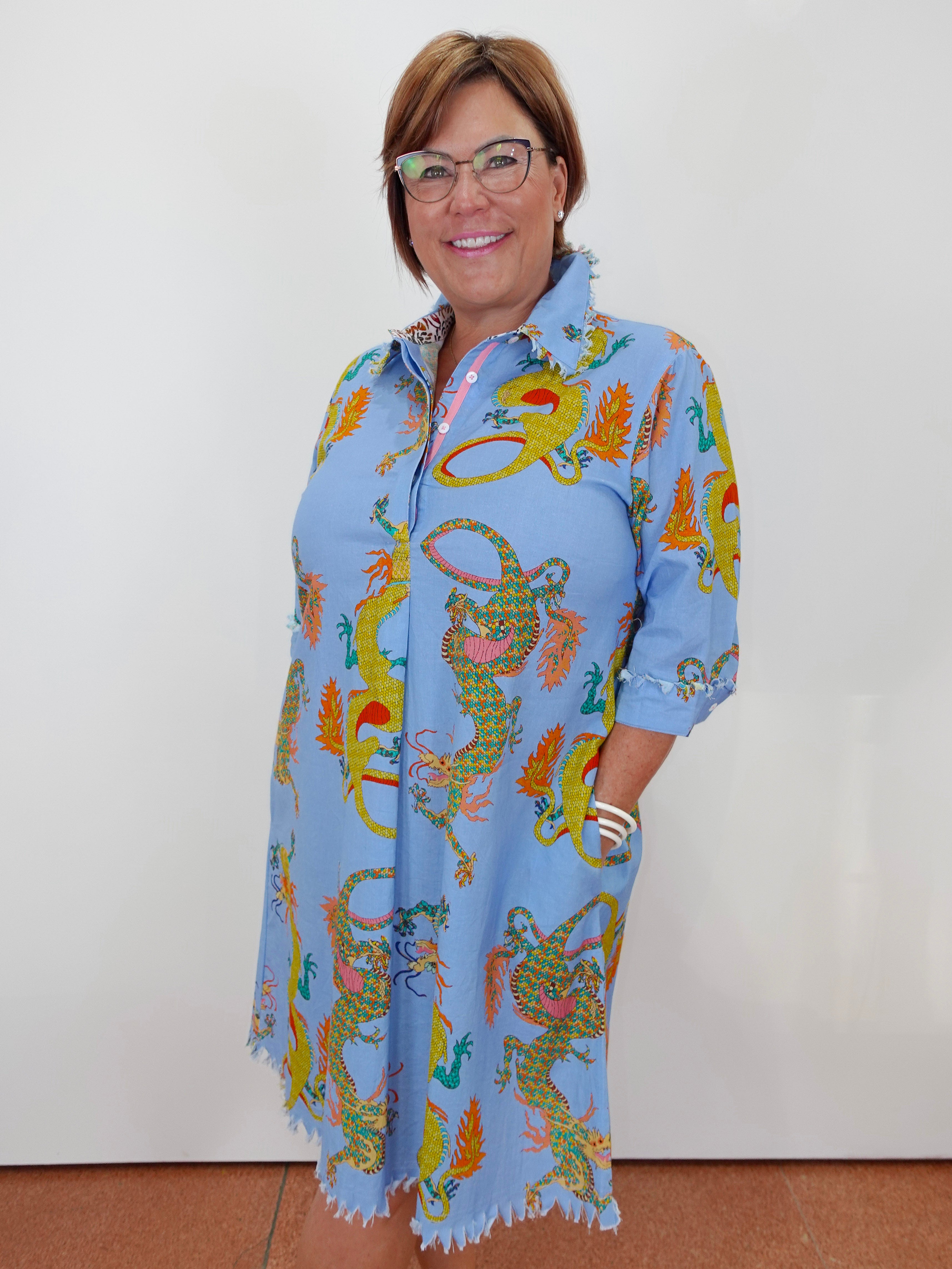 A woman wearing glasses and a light blue collared shirt dress with a colorful dragon print, standing against a plain white wall.