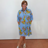 A woman wearing a light blue button-up shirt dress with a colorful, whimsical cartoon-style print, standing against a plain white wall.