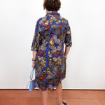 A woman viewed from behind wearing a blue button-up shirt dress with a vibrant butterfly and floral print in red, yellow, and green. She is holding a light blue handbag and standing against a plain white wall on a terracotta-colored floor.