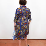 A woman viewed from behind wearing a blue button-up shirt dress with a vibrant butterfly and floral print in red, yellow, and green. She is holding a light blue handbag and standing against a plain white wall on a terracotta-colored floor.