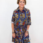 An older woman stands against a plain white wall wearing a short-sleeve button-up shirt dress with a colorful butterfly and floral print in blue, yellow, red, and brown tones. She is holding a light blue handbag.