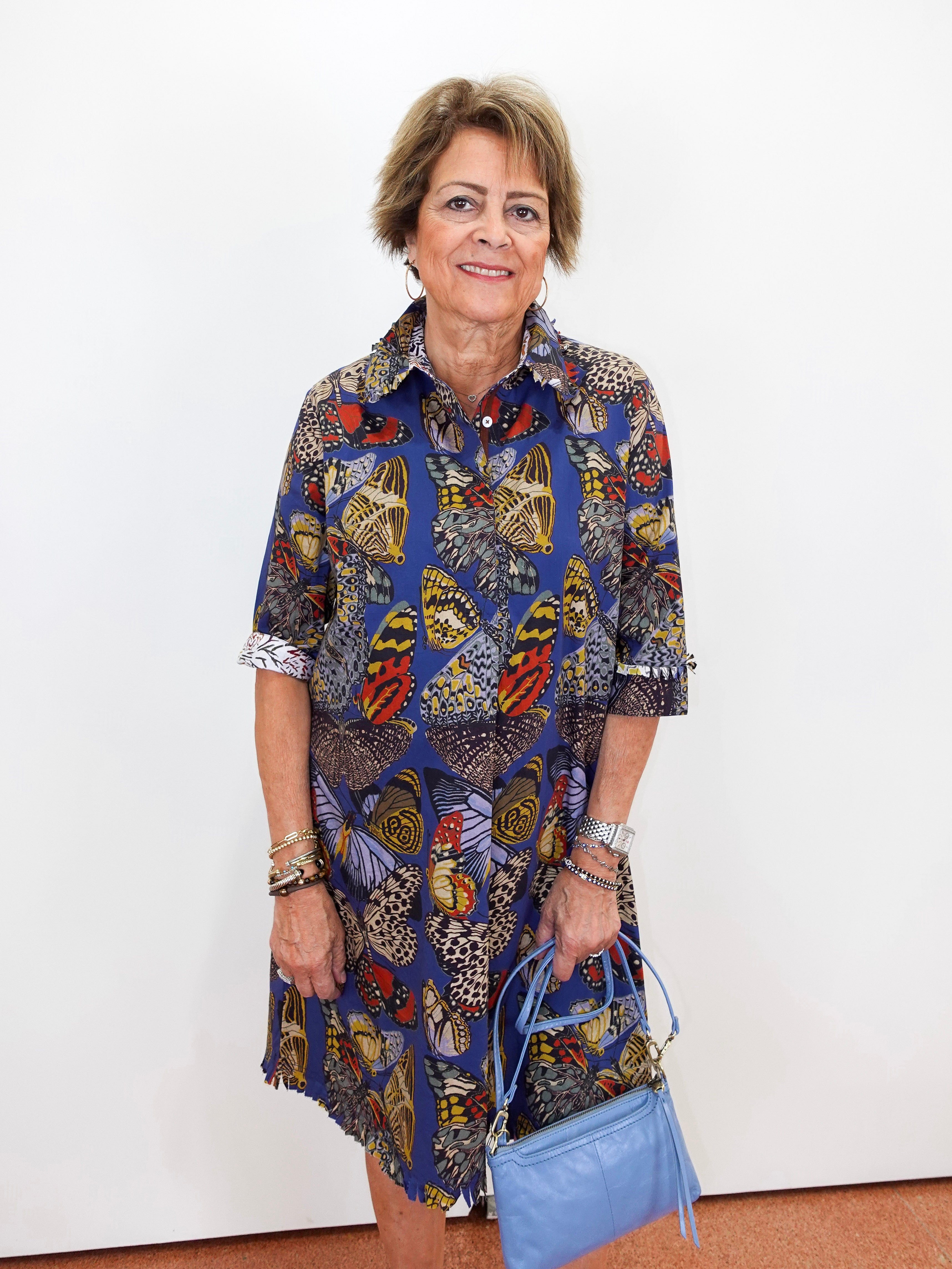 An older woman stands against a plain white wall wearing a short-sleeve button-up shirt dress with a colorful butterfly and floral print in blue, yellow, red, and brown tones. She is holding a light blue handbag.