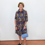 A woman stands against a white wall wearing a short-sleeve button-up shirt dress with a colorful butterfly print in blue, red, yellow, and brown. She is holding a light blue handbag and wearing black slide sandals.