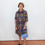 A woman stands against a white wall wearing a short-sleeve button-up shirt dress with a colorful butterfly print in blue, red, yellow, and brown. She is holding a light blue handbag and wearing black slide sandals.