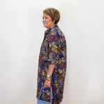 An older woman stands in profile against a plain white wall, wearing a loose, long-sleeve button-up shirt dress with a dense multicolor abstract print in blue, yellow, red, black, and white. She is holding a light blue handbag.