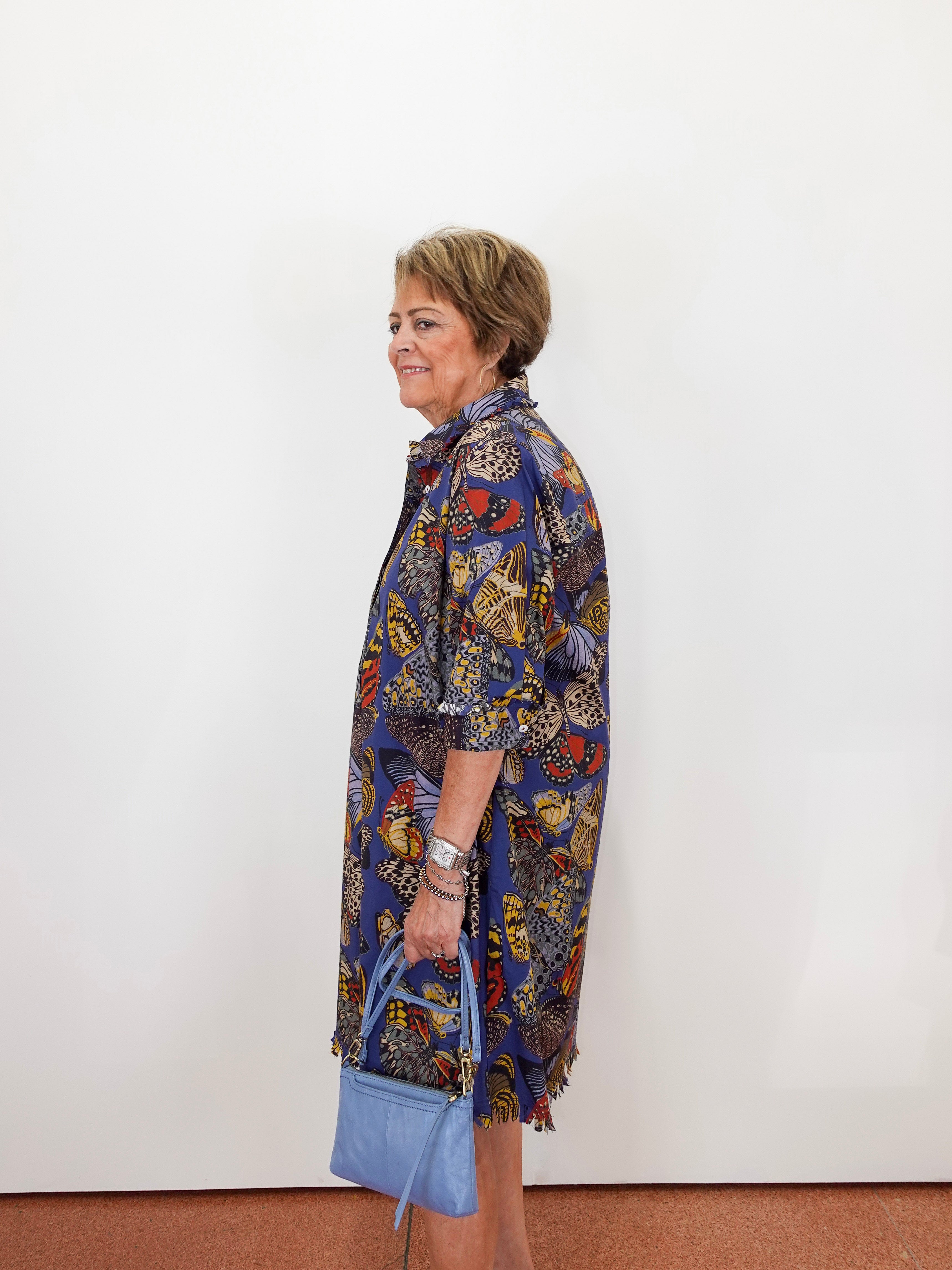 An older woman stands in profile against a plain white wall, wearing a loose, long-sleeve button-up shirt dress with a dense multicolor abstract print in blue, yellow, red, black, and white. She is holding a light blue handbag.