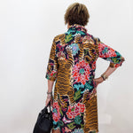 A woman viewed from behind wearing a vibrant, multicolored tiger-and-floral print shirt dress, holding a black handbag against a plain white wall.