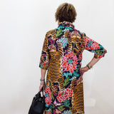 A woman viewed from behind wearing a vibrant, multicolored tiger-and-floral print shirt dress, holding a black handbag against a plain white wall.