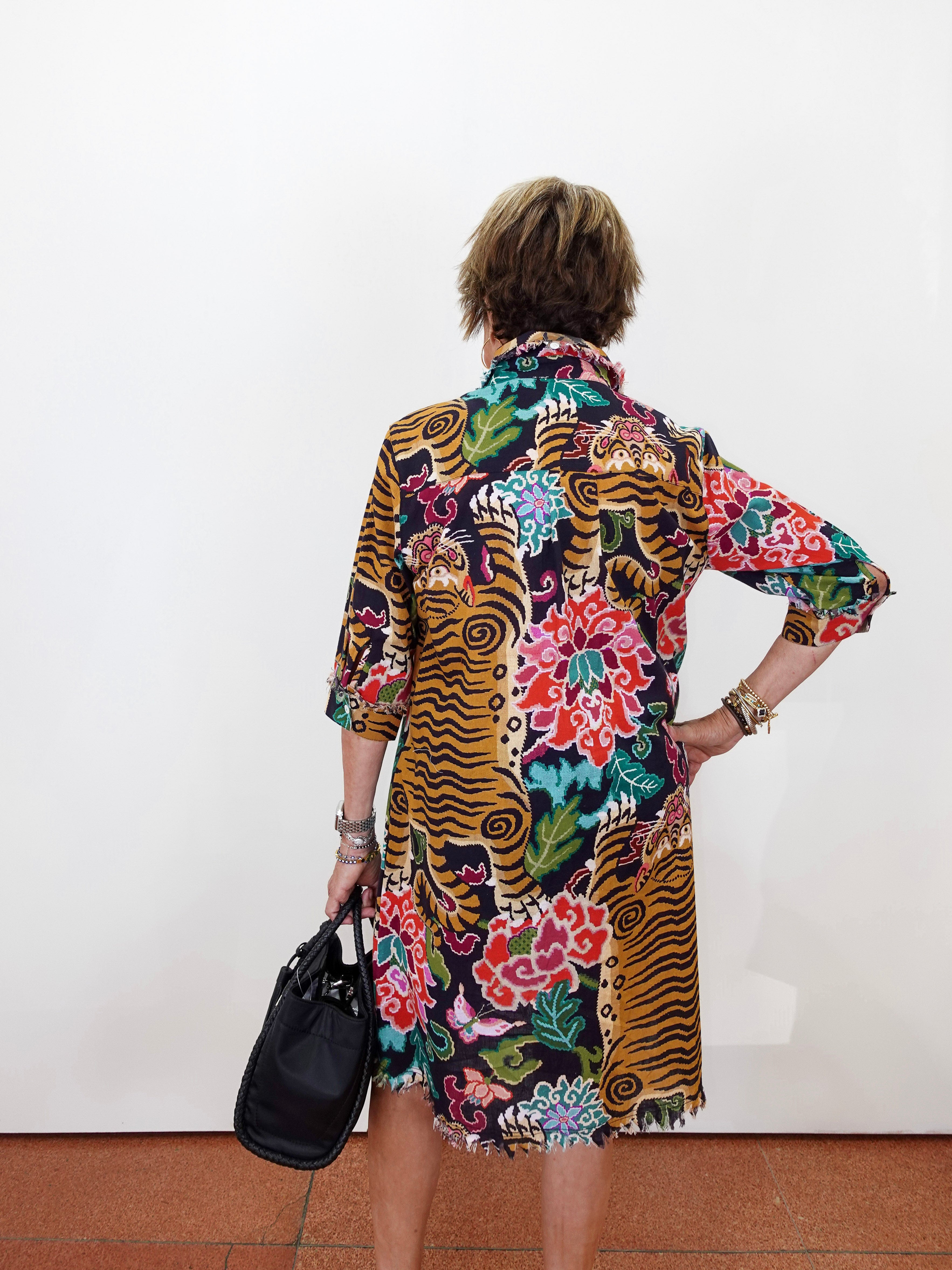 A woman viewed from behind wearing a vibrant, multicolored tiger-and-floral print shirt dress, holding a black handbag against a plain white wall.