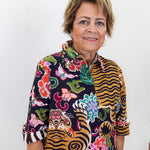 A woman with short blonde hair wearing a colorful, patterned button-up shirt with tiger stripes and floral motifs, standing against a plain white background.