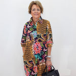 A woman with short blonde hair stands against a white wall wearing a colorful, patterned button-up shirt dress with tiger stripes and floral motifs. She is holding a black handbag.