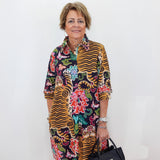 A woman with short blonde hair stands against a white wall wearing a colorful, patterned button-up shirt dress with tiger stripes and floral motifs. She is holding a black handbag.