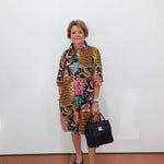 A woman stands against a white wall wearing a colorful mixed-media print shirt dress with tiger stripes and floral patterns, holding a black handbag.