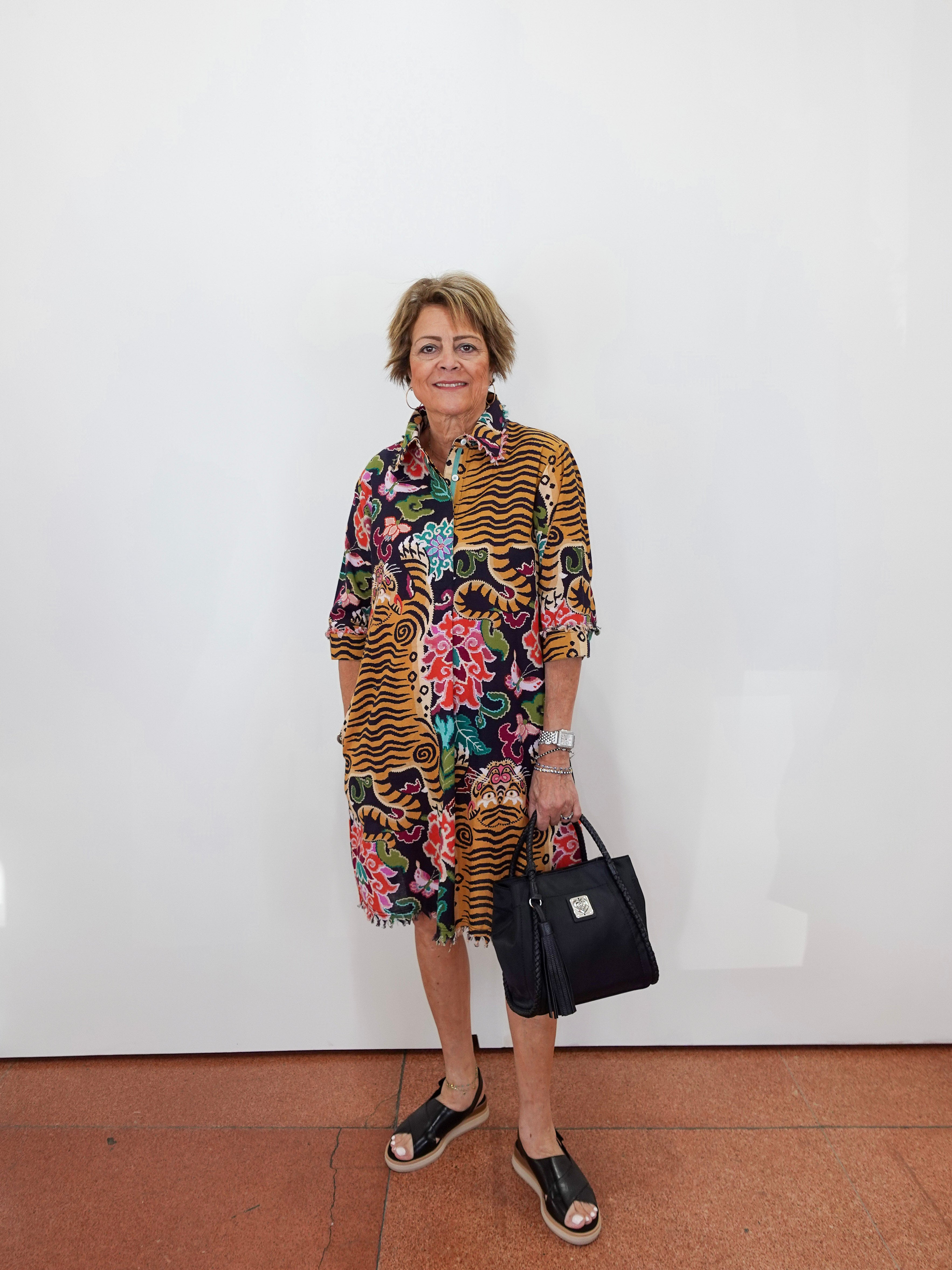 A woman stands against a white wall wearing a colorful mixed-media print shirt dress with tiger stripes and floral patterns, holding a black handbag.