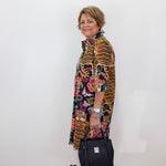 A woman stands in profile against a plain white wall, wearing a long-sleeved, open-front tunic dress with a bold multicolor print featuring tiger stripes, floral motifs, and colorful abstract shapes. She is holding a black handbag and wearing black sandals.
