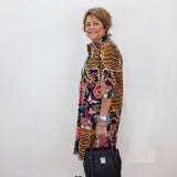 A woman stands in profile against a plain white wall, wearing a long-sleeved, open-front tunic dress with a bold multicolor print featuring tiger stripes, floral motifs, and colorful abstract shapes. She is holding a black handbag and wearing black sandals.